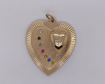 Puede incluir: Colgante dorado en forma de corazón con un corazón más pequeño en el centro. El colgante está adornado con una serie de piedras preciosas de colores y un diamante. El colgante tiene una superficie texturizada y un lazo para una cadena.