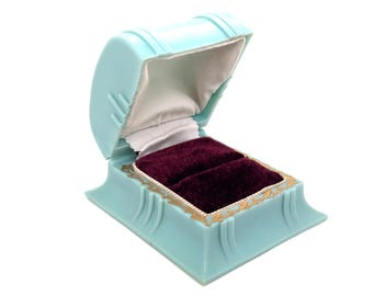 May include: A vintage, open, light blue ring box with a burgundy velvet interior. The box has a gold trim and a white satin lining on the inside of the lid. The box is designed to hold two rings.