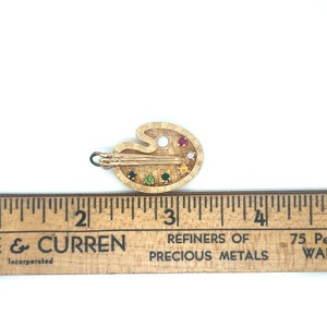 Puede incluir: Un amuleto de paleta de artista de color dorado con un pincel y piedras preciosas de colores. El amuleto mide aproximadamente 2,5 cm de largo.
