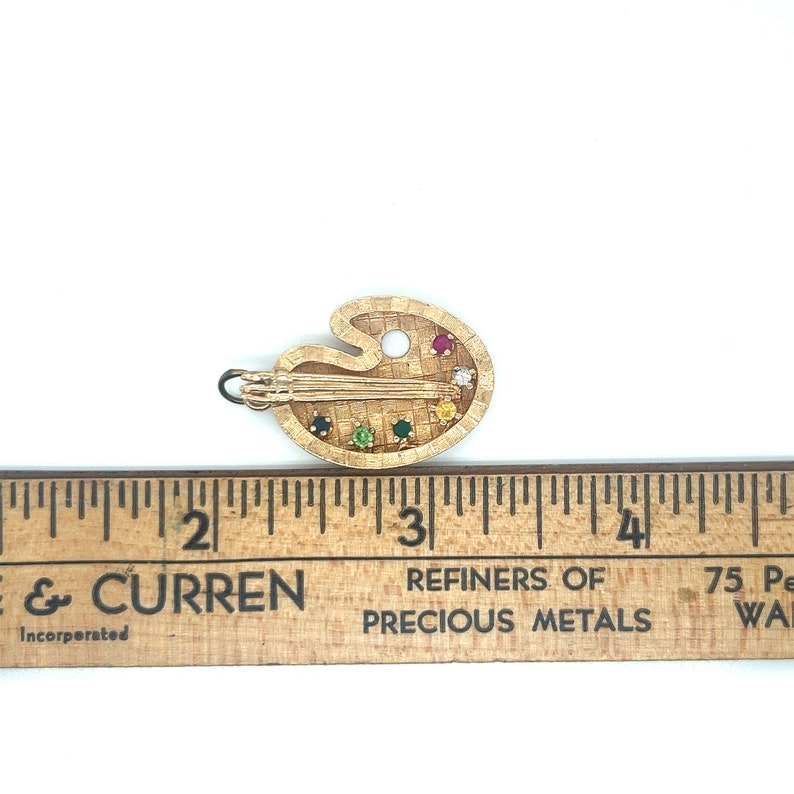 Puede incluir: Un amuleto de paleta de artista de color dorado con un pincel y piedras preciosas de colores. El amuleto mide aproximadamente 2,5 cm de largo.
