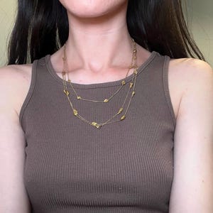 May include: A layered gold-toned necklace with small, irregular-shaped charms. The necklace is worn over a brown ribbed tank top. The jewelry has a delicate appearance and is suitable for everyday wear or special occasions.