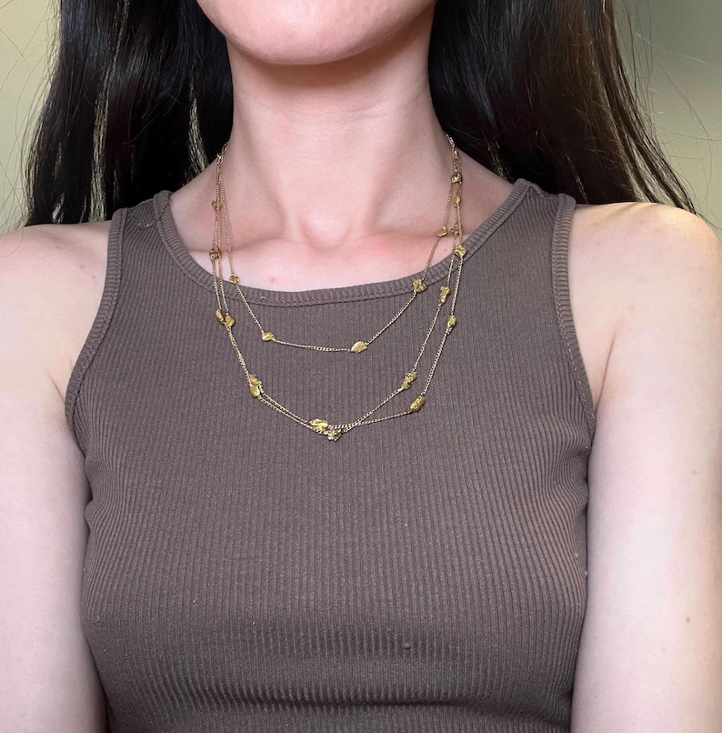 May include: A layered gold-toned necklace with small, irregular-shaped charms. The necklace is worn over a brown ribbed tank top. The jewelry has a delicate appearance and is suitable for everyday wear or special occasions.