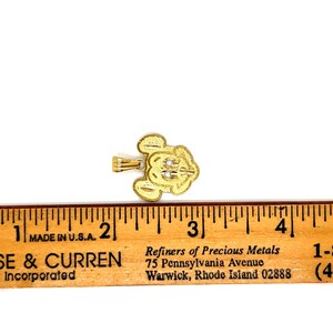 K&ouml;nnte beinhalten: Ein goldfarbener Anh&auml;nger in Form eines Zeichentrickkopfes mit drei kleinen wei&szlig;en Akzenten. Der Anh&auml;nger befindet sich neben einem in Zoll markierten Lineal. Das Lineal lautet "MADE IN U.S.A."