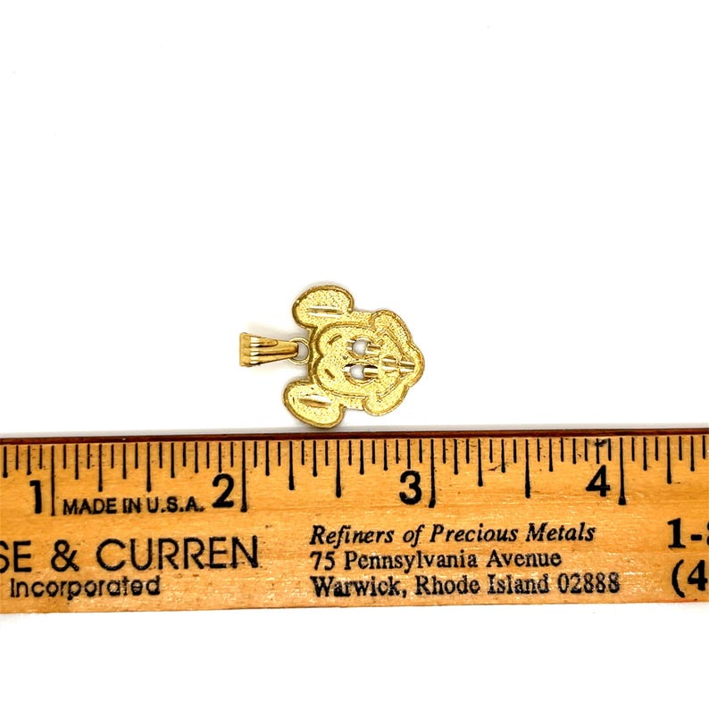 K&ouml;nnte beinhalten: Ein goldfarbener Anh&auml;nger in Form eines Zeichentrickkopfes mit drei kleinen wei&szlig;en Akzenten. Der Anh&auml;nger befindet sich neben einem in Zoll markierten Lineal. Das Lineal lautet "MADE IN U.S.A."