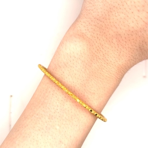 K&ouml;nnte beinhalten: Ein goldfarbenes Armreif-Armband mit strukturiertem Design. Das Armband ist ein d&uuml;nnes, massives Band mit einem sich wiederholenden Muster aus kleinen Kreisen und Linien. Der Schmuck wird am Handgelenk getragen.