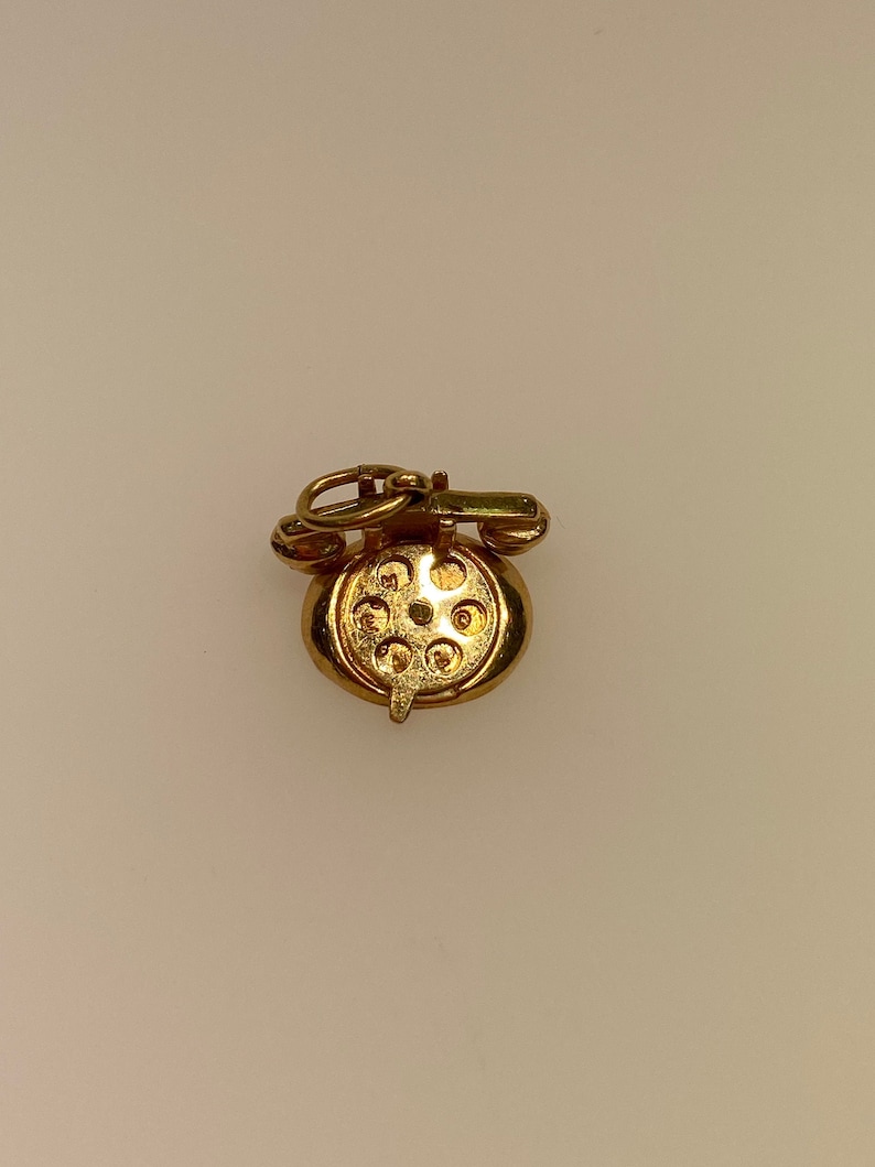 May include: A gold-tone rotary phone charm with a detailed design. The charm has a loop at the top for attaching to a necklace or bracelet.
