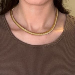 May include: A gold chain necklace with a woven design. The necklace is worn by a person with brown hair and a brown tank top.