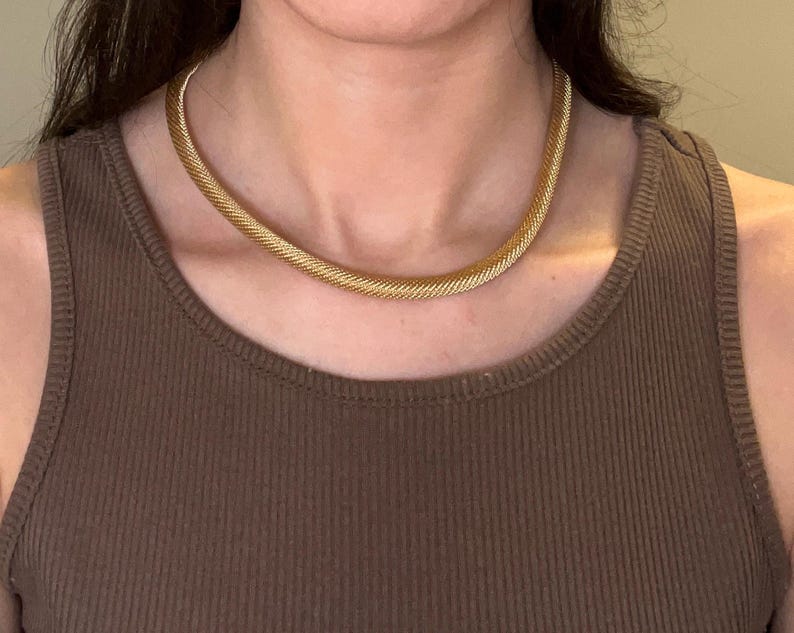 May include: A gold chain necklace with a woven design. The necklace is worn by a person with brown hair and a brown tank top.