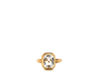 Può includere: Un anello dorato con una grande pietra preziosa trasparente di forma rettangolare. L'anello ha una fascia semplice con dettagli decorativi sui lati. La pietra è incastonata in una lunetta dorata.