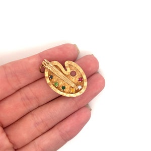 Puede incluir: Un colgante de paleta de artista de color dorado con piedras preciosas de colores. La paleta incluye un pincel y un agujero para una cadena.