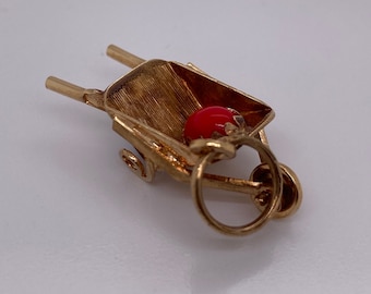 Op de afbeelding: Gouden bedel van een kruiwagen met een rode bal erin. De bedel heeft een lus om aan een ketting of armband te bevestigen.