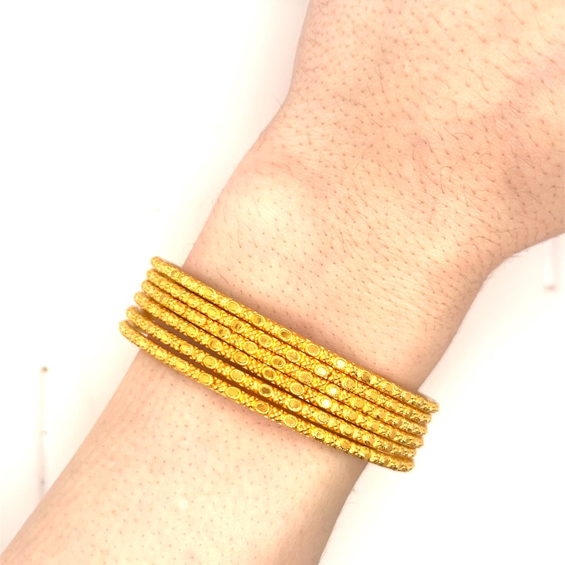 K&ouml;nnte beinhalten: Sechs goldene Armreifen mit aufwendigen Mustern sind an einem Handgelenk gestapelt. Die Armreifen haben eine helle, polierte Oberfl&auml;che, die das Licht reflektiert. Das Design umfasst kleine, sich wiederholende kreisf&ouml;rmige und lineare Details.