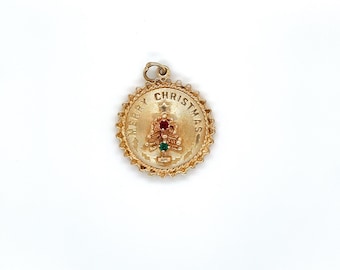 May include: A gold-coloured Christmas charm with a textured edge and the words "MERRY CHRISTMAS" around the perimeter. A small Christmas tree with red and green accents is in the centre. The charm has a loop for attaching to a chain.
