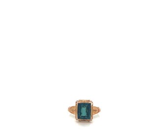 May include: A gold ring featuring a rectangular, emerald-cut teal gemstone set in a prong setting. The band of the ring displays intricate, textured metalwork details on both sides of the central stone, creating a structured and ornate appearance.