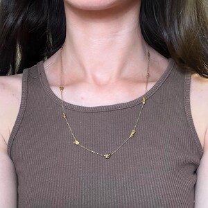 May include: A gold-colored necklace with small, irregularly shaped charms. The necklace is worn over a brown ribbed tank top. The chain is delicate, and the charms are evenly spaced along its length. The overall aesthetic is minimalist and elegant.