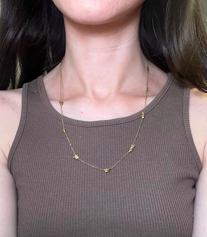 May include: A gold-colored necklace with small, irregularly shaped charms. The necklace is worn over a brown ribbed tank top. The chain is delicate, and the charms are evenly spaced along its length. The overall aesthetic is minimalist and elegant.