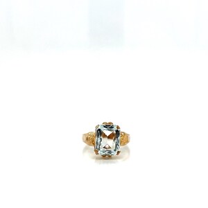 May include: A gold ring featuring a large, rectangular, light blue gemstone. The ring has intricate detailing on the band and around the gemstone setting. The ring is set against a white background.