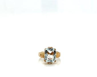 Può includere: Un anello d'oro con una grande pietra preziosa rettangolare azzurra. L'anello presenta dettagli intricati sulla fascia e intorno all'incastonatura della pietra. L'anello è posto su uno sfondo bianco.