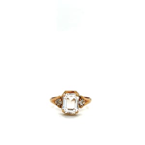 May include: A gold-toned ring featuring a large, rectangular, clear gemstone as the centerpiece. Two smaller, round, clear gemstones flank the main stone. The ring has a classic, elegant design.