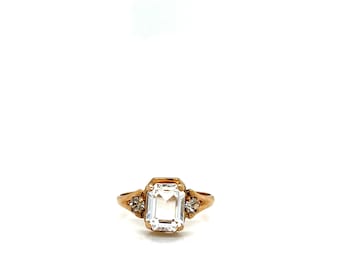 Può includere: Un anello dorato con una grande gemma trasparente rettangolare al centro. Due gemme rotonde più piccole e trasparenti affiancano la pietra principale. L'anello ha un design classico ed elegante.