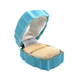 May include: An open, vintage-style, light blue ring box. The box is rectangular with a ribbed design and gold trim. Inside, there are two beige cushions for rings, set against a white satin lining. The box is open, revealing its contents.