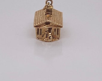 Puede incluir: Un amuleto de oro en forma de una pequeña casa con techo y chimenea.
