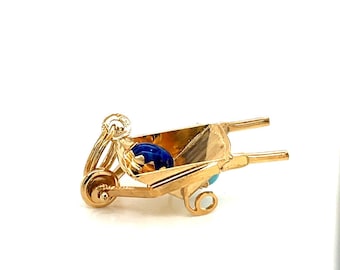 Op de afbeelding: Een gouden bedel in de vorm van een kruiwagen met een blauwe en turquoise steen erin.