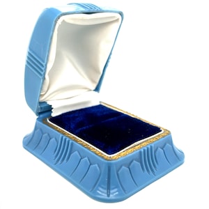 May include: An open, light blue ring box with a white satin interior and a deep blue velvet insert. The box has a gold trim and a decorative, Art Deco-inspired design.