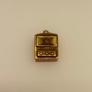 May include: A gold-toned pendant charm in the shape of a small rectangular box with a round, clear glass window and three round, gold-toned buttons.