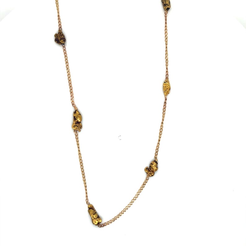 May include: A gold-colored necklace featuring irregularly shaped gold nuggets spaced along a delicate chain. The nuggets have a textured appearance, and the chain is a warm, light gold color. The necklace is displayed against a white background.