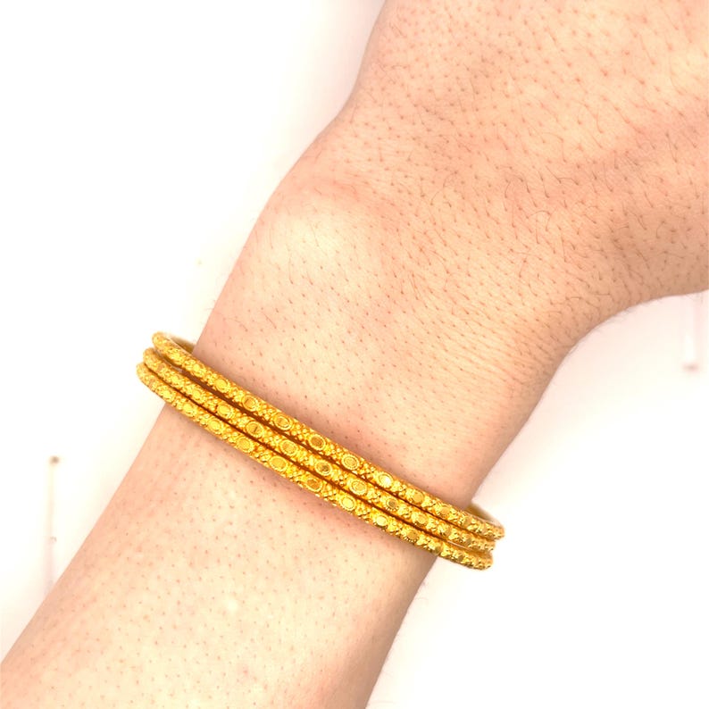 K&ouml;nnte beinhalten: Drei goldene Armreifen mit aufwendigen, sich wiederholenden Mustern. Die Armreifen sind an einem Handgelenk gestapelt und zeigen ihr detailliertes Design und ihre warme, goldene Farbe. Die Armreifen scheinen aus Metall zu sein.