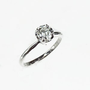 May include: A silver ring with a single round diamond set in a prong setting. The ring is engraved with the text "14K" on the inside of the band.