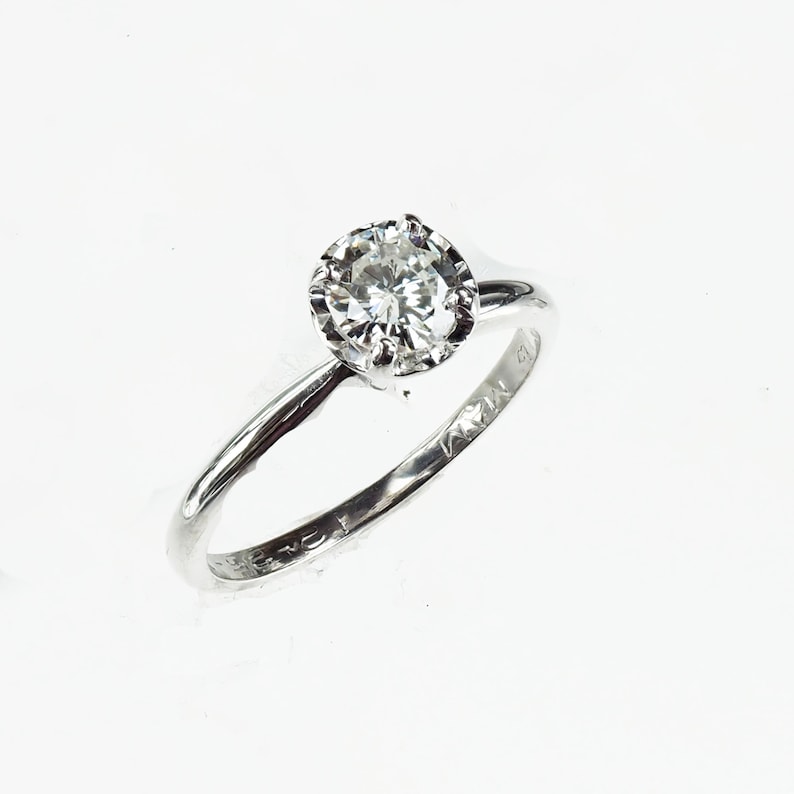 May include: A silver ring with a single round diamond set in a prong setting. The ring is engraved with the text "14K" on the inside of the band.