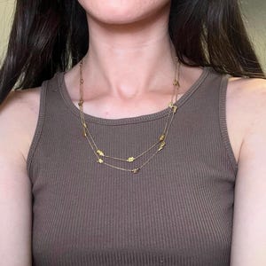 May include: A gold-colored necklace with two strands, adorned with small, leaf-shaped charms. The necklace is worn over a brown ribbed tank top, creating a simple yet elegant look. The jewelry is delicate and adds a touch of sparkle.
