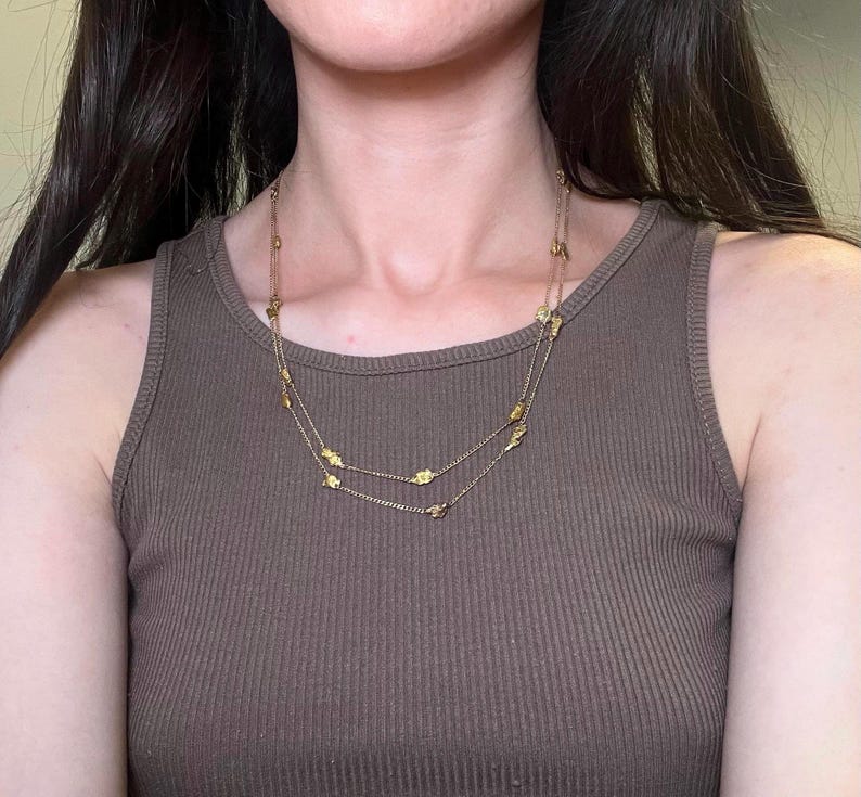 May include: A gold-colored necklace with two strands, adorned with small, leaf-shaped charms. The necklace is worn over a brown ribbed tank top, creating a simple yet elegant look. The jewelry is delicate and adds a touch of sparkle.