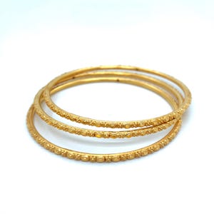 K&ouml;nnte beinhalten: Drei goldene Armreifen mit aufwendigen Mustern. Die Armreifen sind &uuml;bereinander gestapelt und zeigen ihr filigranes Design. Die goldene Farbe verleiht ihnen einen luxuri&ouml;sen Look, ideal, um jedem Outfit einen Hauch von Eleganz zu verleihen.