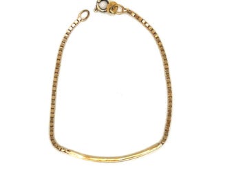 May include: A gold-toned bracelet featuring a box chain design connected to a smooth, curved metal bar. The piece includes a circular spring ring clasp at one end and a small jump ring at the other, creating a continuous, flexible loop structure.