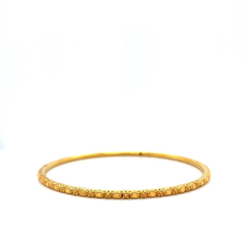 K&ouml;nnte beinhalten: Ein goldener Armreif mit einem sich wiederholenden Muster aus kleinen, erhabenen ovalen Formen. Das Armband hat eine helle, gl&auml;nzende Goldfarbe und ist vor einem wei&szlig;en Hintergrund platziert. Der Schmuck ist schlicht und elegant.