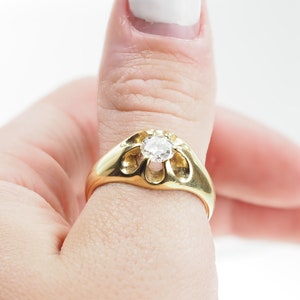 May include: A gold ring featuring a large, clear diamond set in a distinctive, open-work design. The ring is worn on a person's thumb.