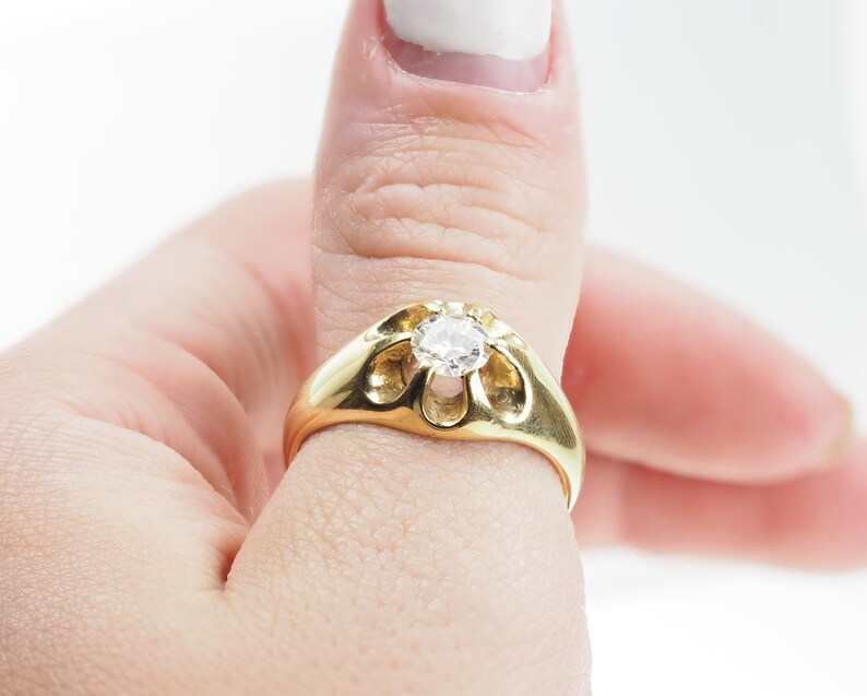 May include: A gold ring featuring a large, clear diamond set in a distinctive, open-work design. The ring is worn on a person's thumb.