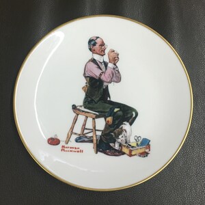 May include: A white ceramic plate with a gold rim features a painting of a man sitting on a stool, sewing. A cat sits beside him. The plate is signed "Norman Rockwell" in red.