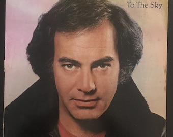 May include: A vintage vinyl record album cover featuring Neil Diamond. The album cover is a close-up of Neil Diamond's face, with a black leather jacket and a red shirt. The album title is "On The Way To The Sky".