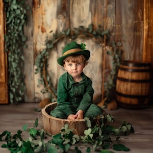 Puede incluir: Un niño pequeño con un sombrero verde y dorado con un trébol en la parte superior se sienta en un barril de madera. El niño lleva un mono verde y está rodeado de hojas verdes.