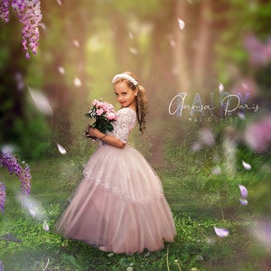 May include: A young person in a pink dress and a floral headband holds a bouquet of pink flowers in a forest setting. The dress has a lace bodice and a layered tulle skirt. Purple wisteria hangs from a tree, and petals float in the air.
