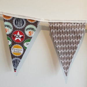 Puede incluir: Una guirnalda de tela marrón y blanca con un tema de soda vintage. La guirnalda presenta una variedad de diseños de tapas de botellas coloridas con texto como "Club Soda", "Cola", "Sarsaparilla" y "Cream Soda".