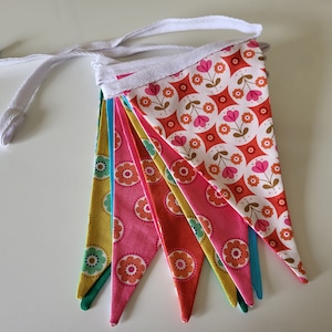 Puede incluir: Una pancarta de tela colorida con una cuerda de algodón blanca. La pancarta presenta un patrón repetido de flores rosas y naranjas sobre un fondo blanco. La pancarta está compuesta por ocho banderas triangulares.