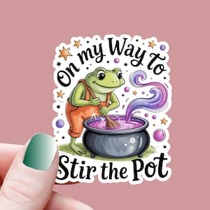 Puede incluir: Una pegatina con una rana de dibujos animados con un mono naranja removiendo una poción púrpura en un caldero negro. El texto dice "On my Way to Stir the Pot" en una fuente caprichosa, con estrellas y burbujas.