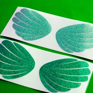 May include: Four green glitter seashell stickers on white sheets of paper. The seashells are arranged in pairs, with two on each sheet.