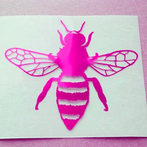 Może przedstawiać: Jasnoróżowa naklejka z pszczołą z detalami skrzydeł i paskowanym odwłokiem. Pszczoła jest wyśrodkowana na białym tle z różowym tłem. Idealna do dodania odrobiny natury.