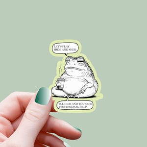 May include: A sticker featuring a cartoon frog with a coffee cup, seated in a meditative pose. The frog has a bored expression and two speech bubbles with the phrases "LET'S PLAY HIDE AND SEEK" and "I'LL HIDE AND YOU SEEK PROFESSIONAL HELP."